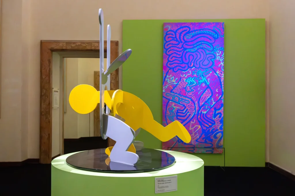 Keith Haring's yellow and white aluminium sculpture Untitled (Head Through Belly), 1986, with a pink and blue painting behind it