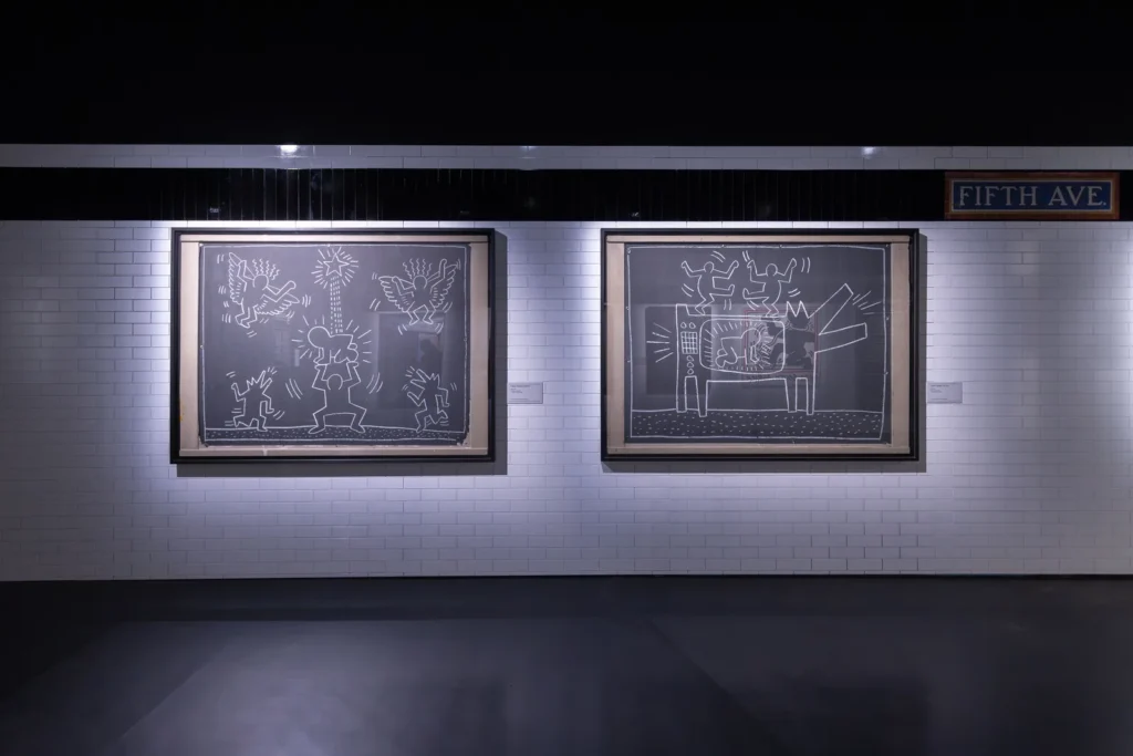 Two Keith Haring Subway Drawings in white chalk on black paper, shown in a room recreating a New York subway station