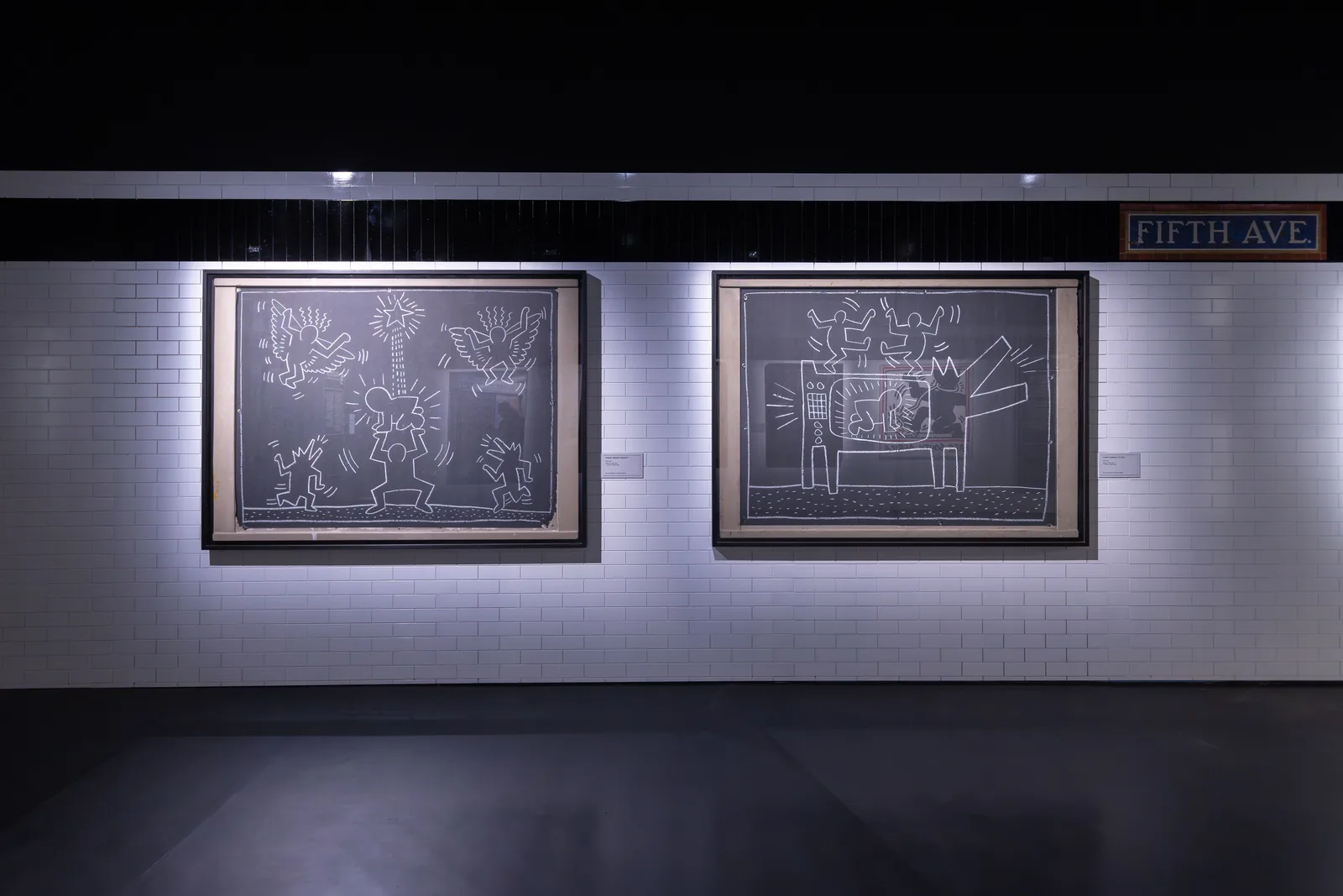 Two Keith Haring Subway Drawings in white chalk on black paper, shown in a room recreating a New York subway station
